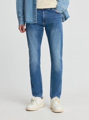 Uomo che indossa jeans blu slim-fit, scarpe da ginnastica bianche, camicia beige e giacca di jeans sbottonata con una mano in tasca.