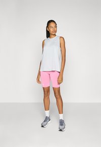 Lys grå ermeløs topp, rosa tettsittende shorts, grå treningssko med hvite detaljer, står mot en enkel lys bakgrunn.