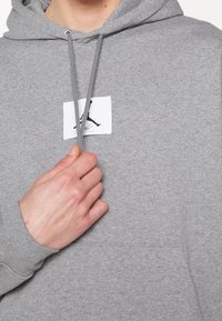 Sweat à capuche gris en tissu doux, avec une poche kangourou frontale et un patch logo carré avec une silhouette noire.