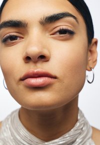 Zbliżenie twarzy o gładkiej, jasnej skórze; wyrazistych brwiach; miękkich, naturalnych ustach; oraz srebrnym, textured szalu. Widoczne małe srebrne kolczyki w kształcie kółek.