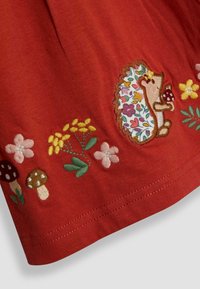 Gelblich-brauner Stoff, der mit einem gestickten Igel, Blumen und Pilzen verziert ist. Das Design umfasst lebendige mehrfarbige Stiche mit detaillierten Mustern.