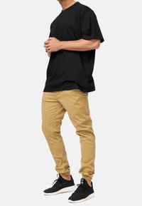 T-shirt en coton noir, coupe décontractée ; pantalon beige fuselé avec poignets élastiques ; baskets noires avec semelles blanches ; modèle debout de profil.