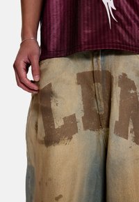 Μακρυμάνικο πουκάμισο βυσσινί με κάθετες ρίγες πάνω από ξεθωριασμένα τζιν σορτς με μεγάλα καφέ γράμματα. Ορατή είναι η φθαρμένη υφή και οι λεκέδες από νερό.