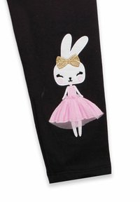 Schwarze Leggings mit einer Applikation eines weißen Kaninchens mit einer goldenen Schleife, das ein pinkes Tüllkleid trägt, auf dem linken Bein.