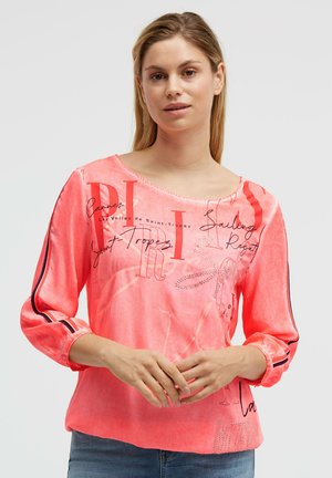 Frau trägt ein korallenrosafarbenes langärmeliges Shirt mit schwarzen und roten Streifen auf den Ärmeln und einem schwarzen, handschriftlich gestalteten Textdesign.
