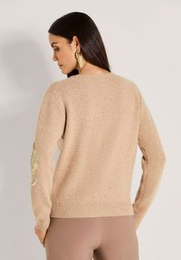 Frau mit langen dunklen Haaren, die einen beigen Strickpullover mit goldener floraler Stickerei an den Ärmeln und hellbraunen Hosen trägt, steht mit dem Rücken zur Kamera.