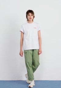 T-shirt en coton blanc avec des manches retroussées et un logo rouge sur la poitrine, associé à un pantalon cargo vert ample et des baskets blanches.