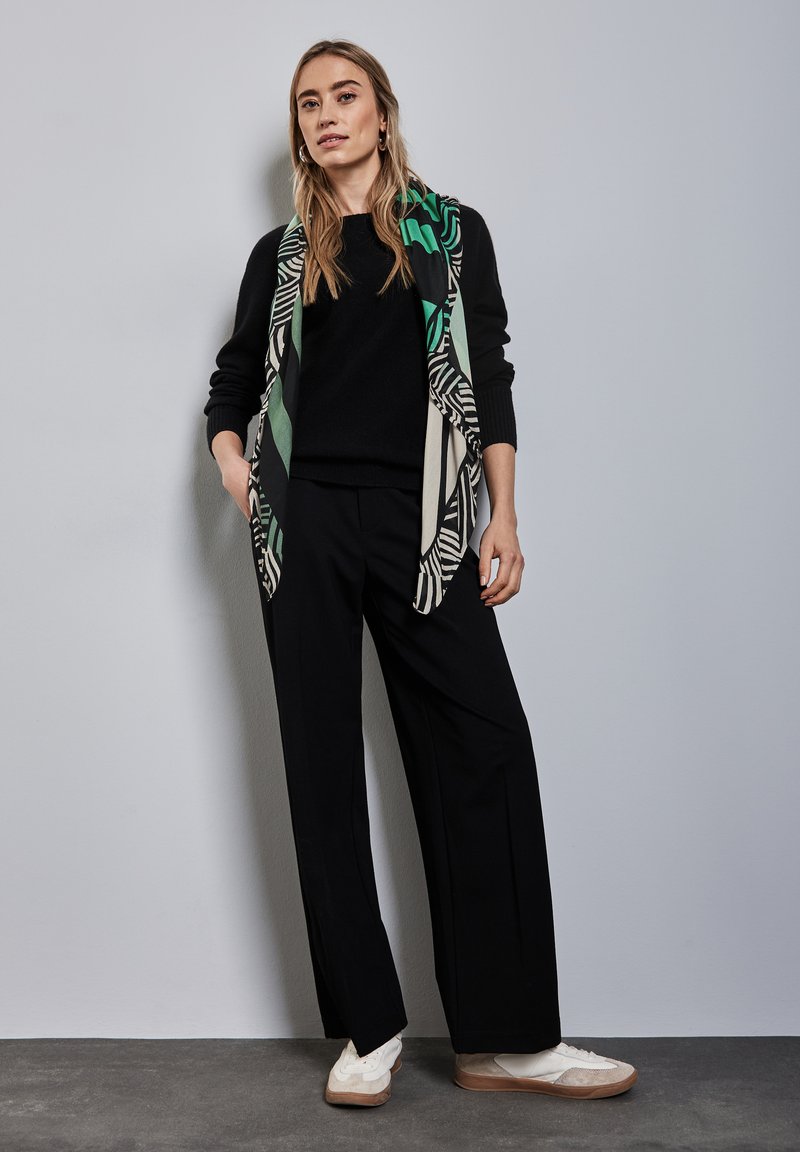 Pull noir et pantalon large noir associés à une écharpe à motifs avec des tons vert et noir. Le modèle se tient sur un sol gris contre un mur clair.
