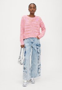 Sieviete rozā adītā džemperī un gaiši zilos plaša silueta darba džinsos, rokās turot mazu sudraba krāsas somiņu, stāvot pret baltu fonu.