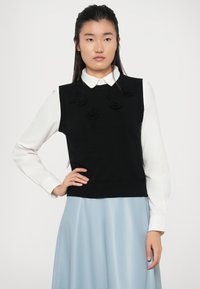 Un pull sans manches noir en tricot avec des détails floraux texturés sur le devant, porté par-dessus une chemise blanche à col, associé à une jupe bleu clair.