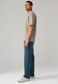 Tricou bej cu mânecă scurtă, cu o croială relaxată, asortat cu blugi negri. Modelul poartă adidași albi, stând în profil pe un fundal neutru.