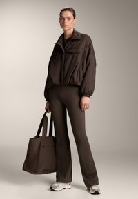 Brown puffer jacket with front pockets, paired with fitted brown leggings and a large brown tote bag. Model wears gray and white athletic shoes.