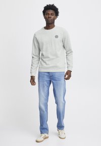  Lichtgrijze sweatshirt met ronde hals en geborduurd logo, gecombineerd met blauwe jeans en witte sneakers met bruine accenten.