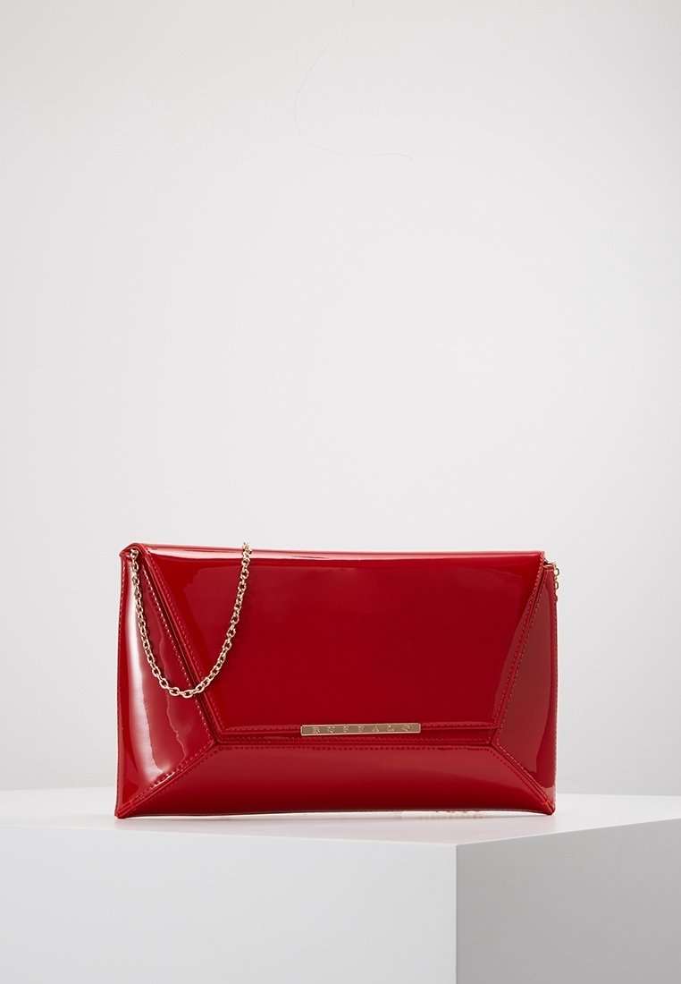 Pochette en cuir verni rouge brillant avec coutures triangulaires et petite bandoulière chaîne dorée, reposant sur une surface blanche.