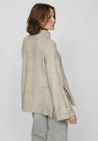 Pull à col roulé en tricot beige avec une coupe ample, épaules tombantes, poignets côtelés et fentes sur les côtés, associé à un pantalon gris.