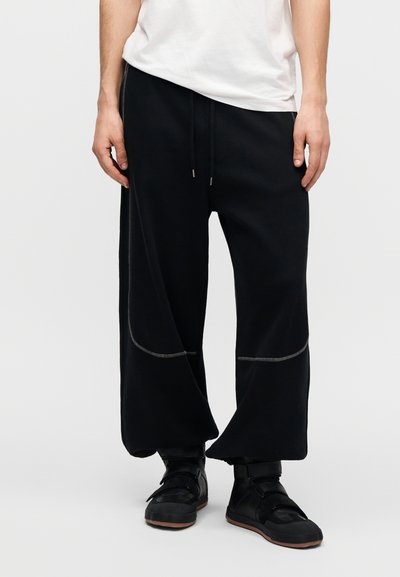 Μαύρο φαρδύ παντελόνι φόρμας με λάστιχο στη μέση και κορδόνι, με αντίθετες γκρι ραφές και άνετη εφαρμογή. Φορεμένο με μαύρα αθλητικά παπούτσια.