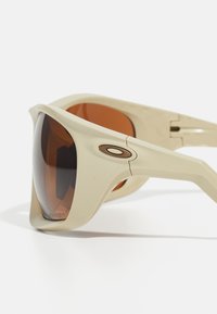 Beige innhyllende solbriller med brune linser. Har en fremtredende logo og robuste rammer, designet for holdbarhet og UV-beskyttelse.