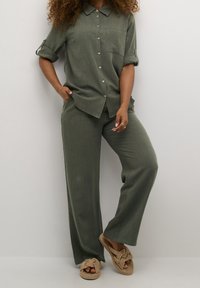 Donna che indossa una camicia verde oliva con bottoni e maniche arrotolate, pantaloni abbinati, in piedi con una mano in tasca e sandali di corda beige.