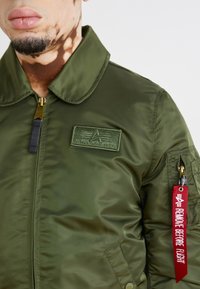 Zelena bomber jakna iz gladkega najlona z zadrgo, rebrastim ovratnikom, obližnim logotipom in rdečo etiketo z napisom "REMOVE BEFORE FLIGHT".