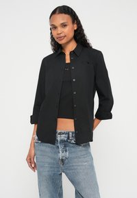 Melna pogāžama krekla izgatavota no gludas auduma ar smailu apkakli, uz krūšukurvja redzama neliela logotipa uzlīme un uzrokotām piedurknēm.