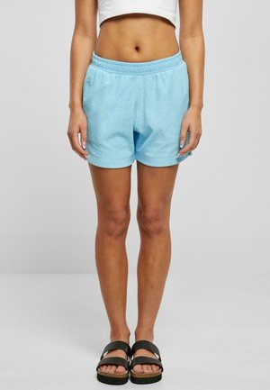 Kvinde iført lyseblå terry-kludsshorts, hvid cropped top og sorte sandaler med dobbelt rem, stående mod en neutral baggrund.