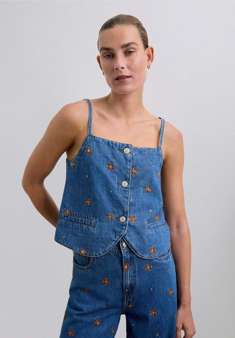 Denim cropped vest met verstelbare dunne bandjes, knoopsluitingen aan de voorkant en florale borduursels in oranje en wit op een blauwe achtergrond.