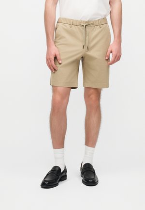 Άνδρας που φοράει μπεζ σορτς με κορδόνι, άσπρες κάλτσες και μαύρα δερμάτινα μοκασίνια, στέκεται μπροστά σε απλό λευκό φόντο.