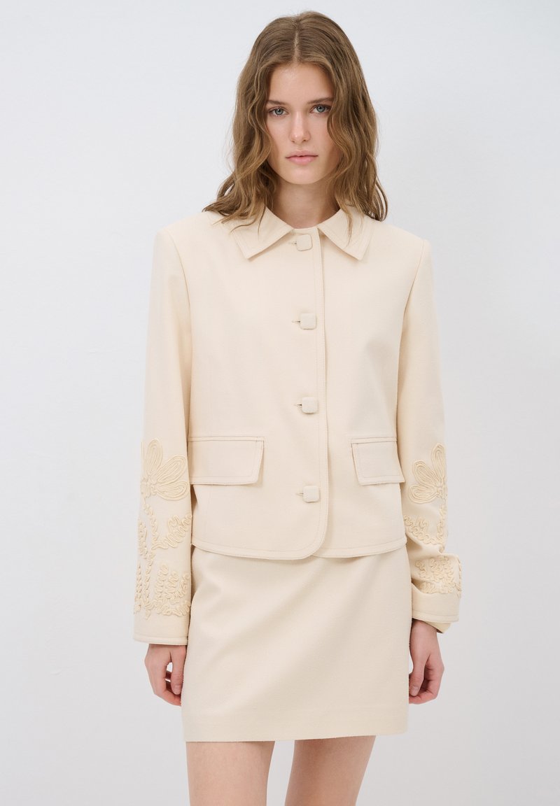 Beige op maat gemaakte jacket met een kraag, vijf knopen en bloemenborduursel op de mouwen; gecombineerd met een bijpassende beige rok.