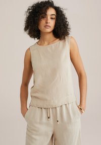 Top sans manches beige en tissu léger, présentant une texture lisse, un col rond et une coupe décontractée. Associé à un pantalon assorti.