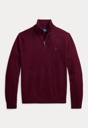 Mann trägt einen burgunderfarbenen Viertel-zip-Pullover über einem gestreiften Hemd, dunkelgrüne Cordhosen, braune Schuhe und eine Armbanduhr, steht vor einem einfarbigen Hintergrund.