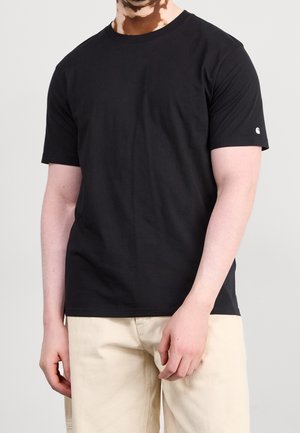 Mand iført en ensfarvet sort kortærmet T-shirt og beige bukser, stående foran en ensfarvet lys grå baggrund.