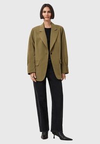 Blazer oversize verde oliva con revers a punta e bottoni neri, abbinato a jeans neri a gamba dritta e décolleté nere a punta.
