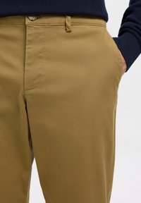Mies, jolla on päällä beige housut, käsi taskussa ja tummansininen neule, joka näkyy vyötäröllä ja ranteessa.