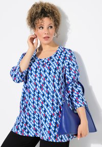Tunica blu con un motivo geometrico in rosso e bianco, scollatura tonda, maniche a tre quarti; abbinata a una borsa triangolare blu.