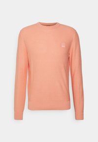 Pull en maille rose clair à col rond, avec poignets et ourlet côtelés, arborant un petit patch logo sur la poitrine. Coupe classique et design texturé.