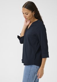 Blusa blu navy, con texture, scollo a V e maniche tre quarti, rifinita con dettagli arricciati sui polsini. Indossata con jeans blu chiaro.