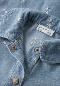 Lyseblå denimskjorte med blomsterbroderi, med klassisk krage, kobberfargede knapper og en vevd vaskeanvisning nær halslinningen.