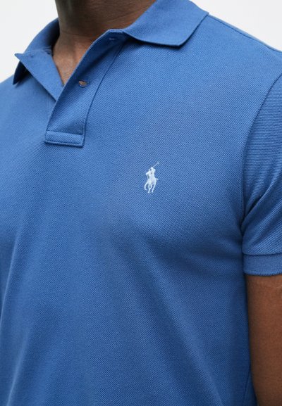 Μπλε πόλο μπλούζα από υφασματοποιημένο ύφασμα, με κλασικό γιακά και κοντά μανίκια. Ένα λευκό λογότυπο είναι κεντημένο στο αριστερό στήθος.
