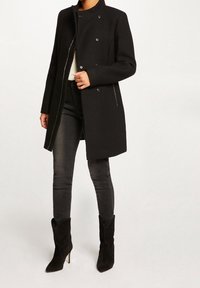 Zwarte getailleerde jas met een hoge kraag, dubbele knoopsluiting en zijritsen. Gecombineerd met donkere skinny jeans en laarzen met hak.