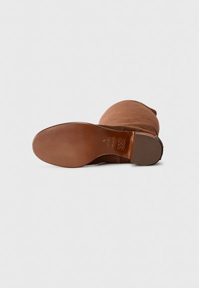 Bota de ante marrón con puntera redondeada, suela de cuero suave y detalles de hardware mínimos. Textura suave con un eje estructurado.