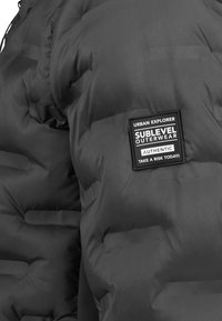 Schwarze gepolsterte Jacke mit einem quadratischen Aufnäher auf dem Ärmel, auf dem steht: "Urban Explorer Sublevel Outerwear Authentisch Nimm heute ein Risiko!"