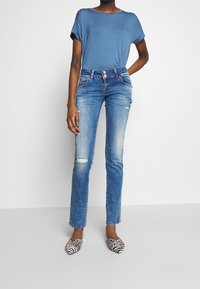 Top de manga corta azul combinado con jeans ajustados de cintura baja y rotos en un tono azul claro, y zapatillas de slip-on en blanco y negro con estampado. Atuendo simple y casual.