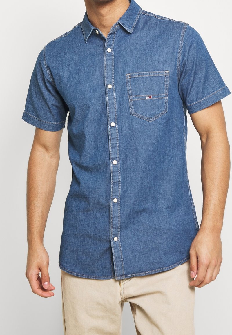 Chemise en jean manches courtes bleu moyen avec un col, une poche avant et des boutons blancs ; présente une texture subtile et une coupe décontractée.