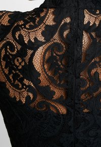 Le tissu en dentelle noire présente des motifs floraux complexes avec une finition texturée. Le design met en avant un détail de fermeture éclair dans le dos et un contraste de peau.