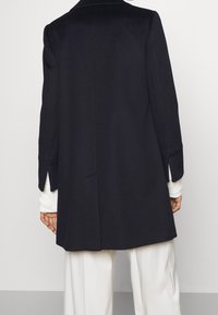 Black wool-blend coat with a tailored fit, notched collar, and side slits. The back features a clean design, paired with a textured white sweater.