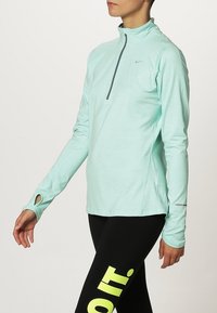 Osoba ubrana w miętowo-zieloną sportową bluzę z długim rękawem i krótkim zamkiem oraz czarne legginsy z jaskrawo żółto-zielonym napisem na udzie.