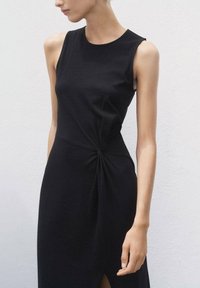 Zwart mouwloos jurkje met een aansluitend ontwerp, voorzien van een gedraaide knoopdetail bij de taille en een zijsplit, gemaakt van een zachte stof.