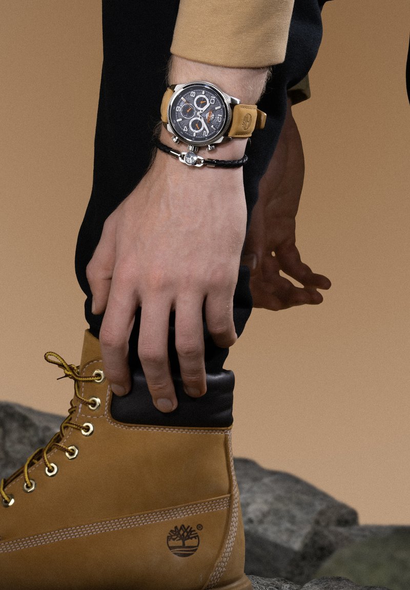 Stivale in pelle marrone con accessori dorati e dettagli neri, abbinato a un orologio cronografo argento con quadrante nero e cinturino beige.