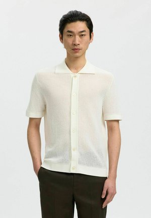 Mann trägt ein kurzärmeliges, cremefarbenes Strickhemd mit Knöpfen und Kragen und dunkle Hose vor einfachem Hintergrund.