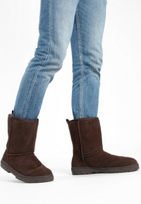 Botas de ante marrón de altura media hasta la pantorrilla, con una abertura amplia y suelas de goma, combinadas con vaqueros azules ajustados. Textura suave y detalles mínimos.